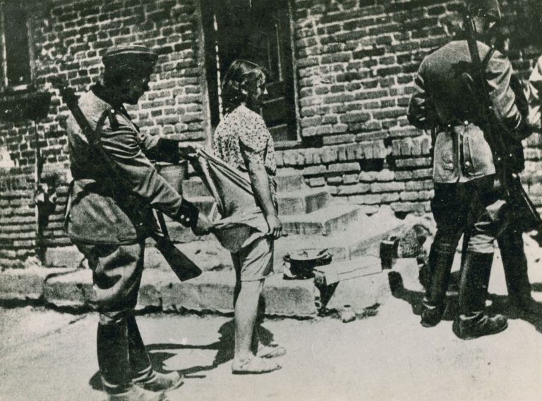 Oborožen nacistični vojak preiskuje žensko, tako da ji sname spodnje perilo, Poljska, okoli 1939-1944. Fotografija: USHMM #73719, avtor Moshe Berry